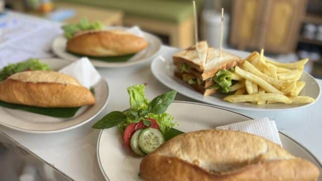 Four plates with sandwiches, one featuring fries and a club sandwich, inspired by Hoi An, garnished with fresh vegetables and herbs.