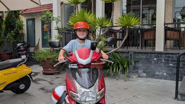 A person wearing an orange helmet sits on a red motorbike in front of a modern Hoi An building with plants.