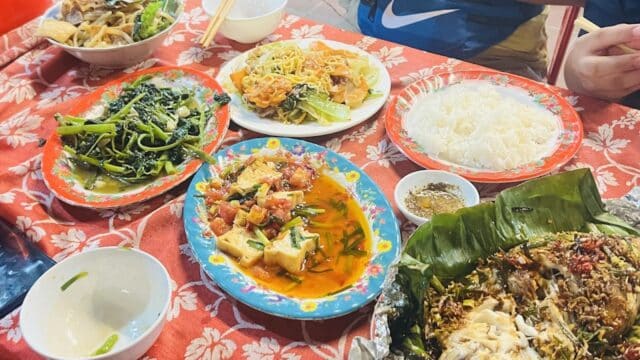 A colorful table of Hoi An Vietnamese dishes with rice, vegetables, and fish on a red floral tablecloth.