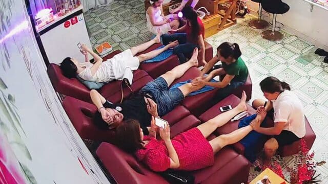 Six people relax on red couches getting foot massages, while two others work in a brightly lit Hoi An spa.