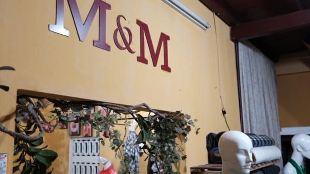 A room in Hoi An with mannequins, fabric rolls, and an “M & M” sign on a yellow wall above a mirror with greenery.