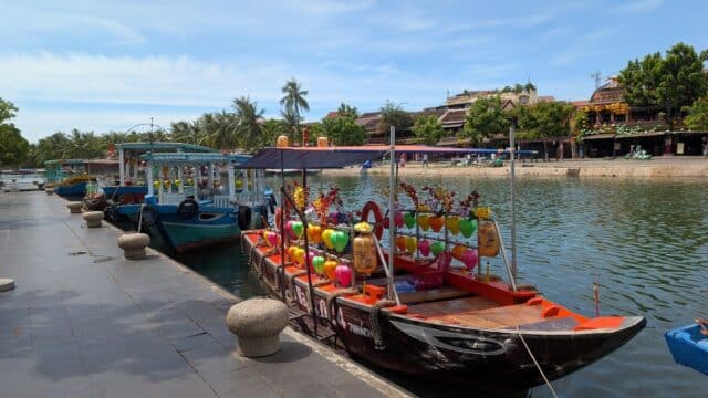Boats decorated with colorful lanterns are docked along Hoi An’s riverside promenade under a bright, sunny sky.