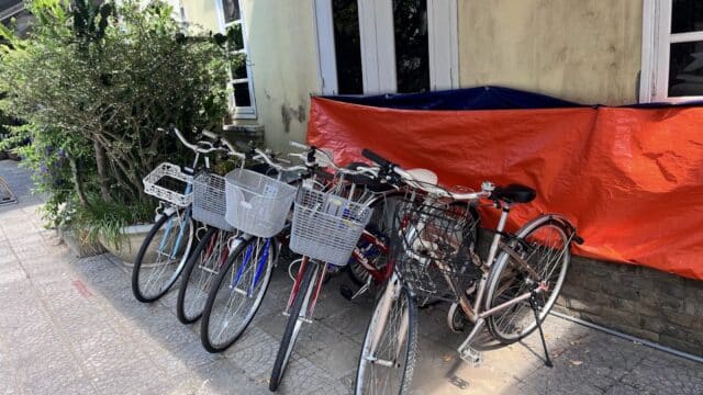 Five bicycles with baskets are parked on a sidewalk in Hoi An beside a building covered with orange tarp.
