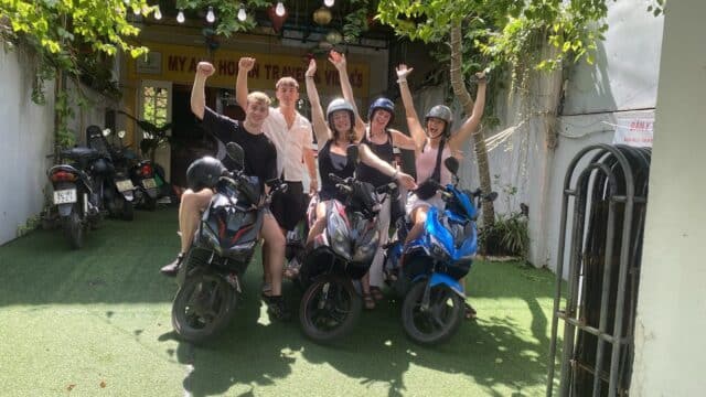 Five people wearing helmets sit on scooters, smiling and raising their arms outside a Hoi An travel agency with greenery overhead.