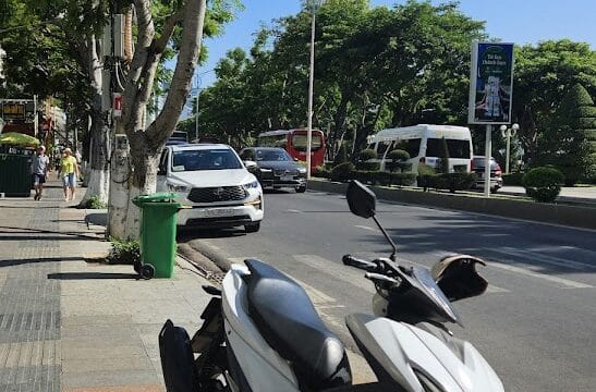A white scooter is parked on a sunny Hoi An sidewalk beside a busy street lined with trees and cars.