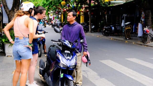 Two women talk to a man standing by a parked scooter in Hoi An, where the street is lined with trees and charming shops.