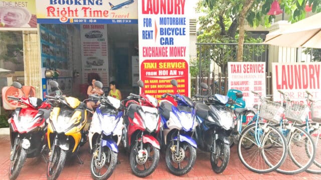 Row of motorbikes and bicycles for rent outside a travel and laundry shop in Hoi An with multiple service signs.