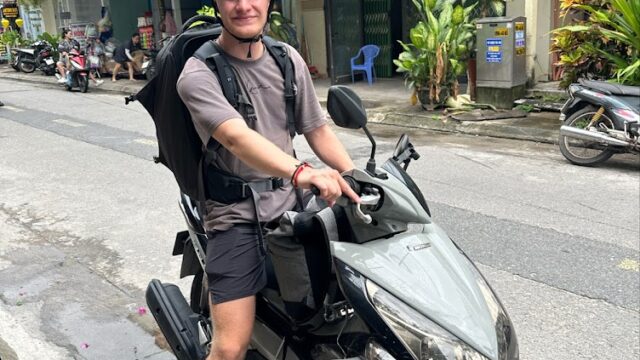 A person wearing a helmet and backpack sits on a gray scooter parked on a street in Hoi An’s urban area.