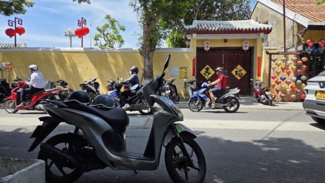 Scooters line the Hoi An street, with people on motorcycles, red lanterns, and a yellow-walled building in the background.