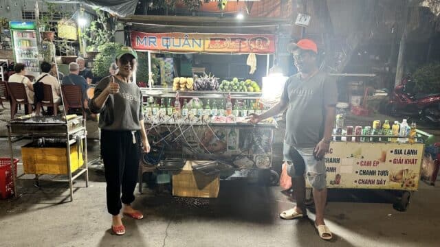 Two people stand smiling at a brightly lit Hoi An street food stall with drinks, fruits, and signs at night.
