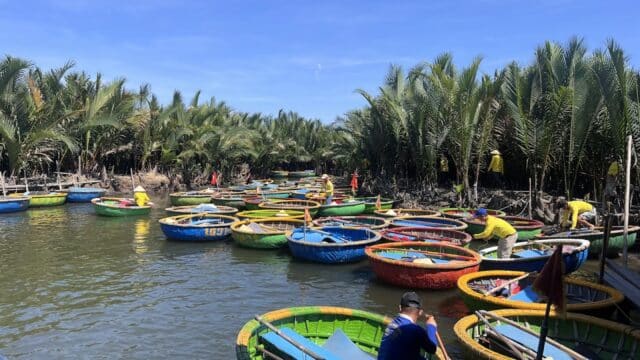 Colorful round boats float on a Hoi An river lined with palm trees, people in yellow shirts aboard and along the banks.
