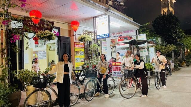 Five people with bicycles smile outside a brightly lit Hoi An building adorned with red lanterns at night.