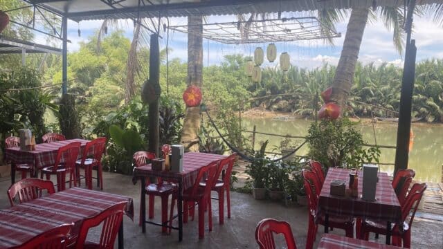 Open-air restaurant in Hoi An with red chairs, plaid tablecloths, and a river view surrounded by lush green trees.