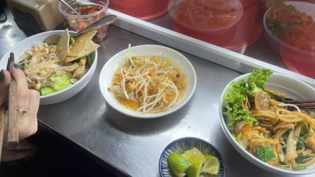 Three bowls of Hoi An Vietnamese noodle dishes with vegetables and lime wedges on a metal countertop.