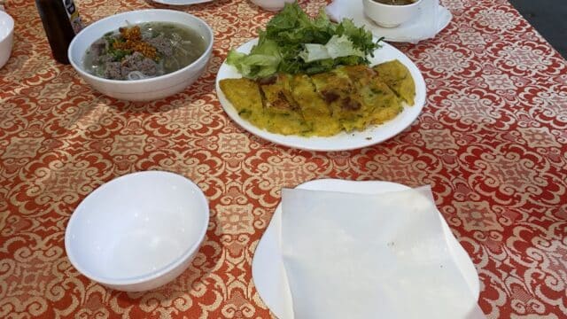 Vietnamese dishes from Hoi An on a red patterned tablecloth, featuring banh xeo, greens, soup, sauce, and empty bowls.