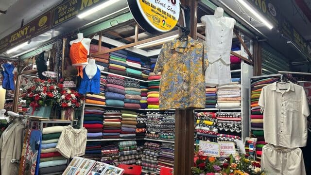 Hoi An market stall with colorful fabric rolls, mannequins in shirts and pants, flowers, and a sign reading Ms. NÔ.