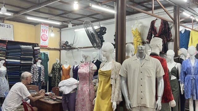 Hoi An clothing shop with mannequins in dresses and shirts, a man at a desk, and fabric rolls lining the shelves.
