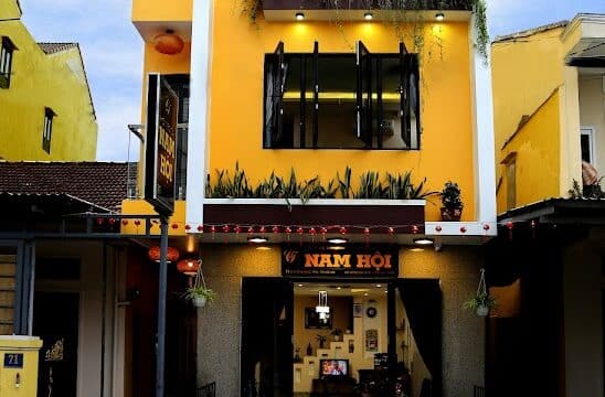 A three-story yellow building with balconies, large windows, and plants in Hoi An, set against a bright blue sky.