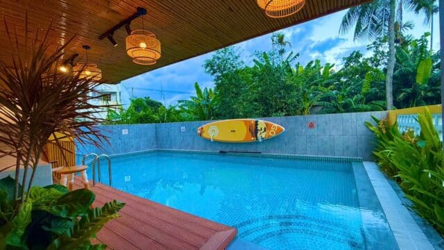 A modern outdoor pool in Hoi An with wooden deck, hanging lights, lush plants, and a yellow paddleboard mounted on a gray wall.