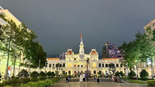 People gather in front of a brightly lit, historic Hoi An building with a flag on top, surrounded by trees at night.