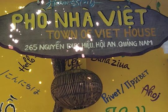 A wooden sign for Pho-Nha Viet House hangs on a colorful wall in Hoi An, with greetings in various languages.