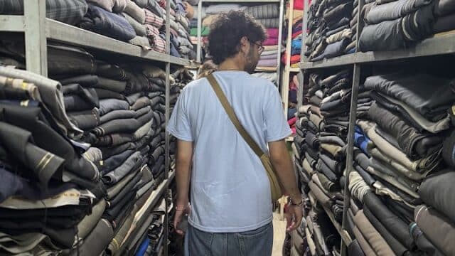 A man stands barefoot in an aisle between shelves stacked with folded fabrics in a brightly lit Hoi An store.