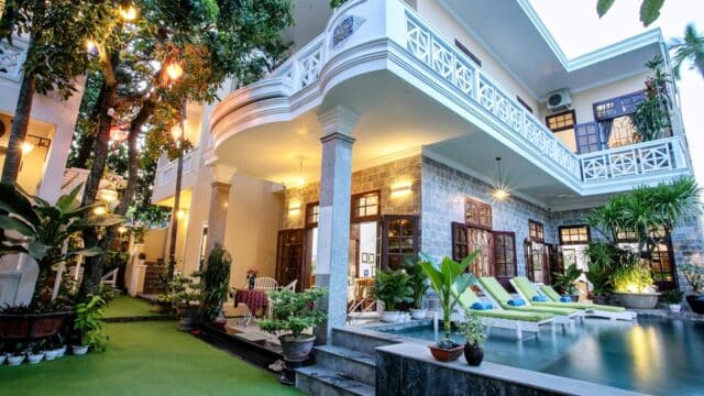 Modern Hoi An villa with balconies, a pool, lounge chairs, lush plants, and warm outdoor lighting in the evening.
