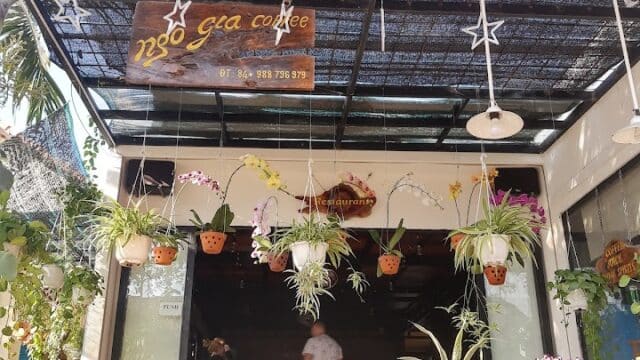Outdoor view of a Hoi An café with hanging plants, a wooden sign, and people sitting inside under a shaded roof.