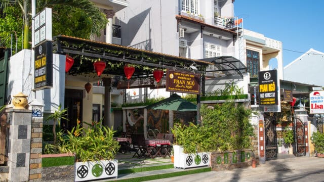 Street view of a Hoi An cafe with outdoor seating, green plants, and red lanterns under a canopy.