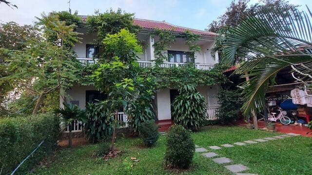 Two-story white house with red roof, draped in green vines, nestled among trees and grass, evokes the charm of Hoi An.