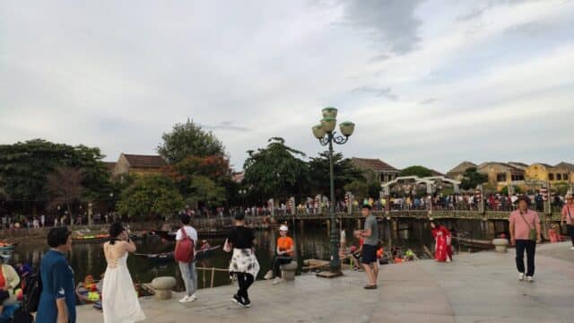 People stroll along Hoi An's riverside promenade with boats and a bridge in the background under a cloudy sky.