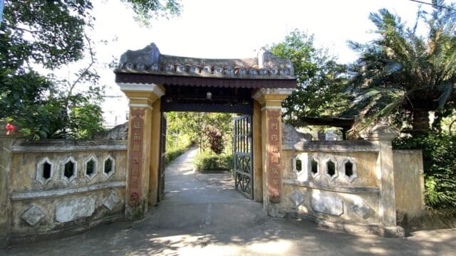 Traditional stone gate with red pillars and yellow accents, opening to a lush Hoi An garden path on a sunny day.