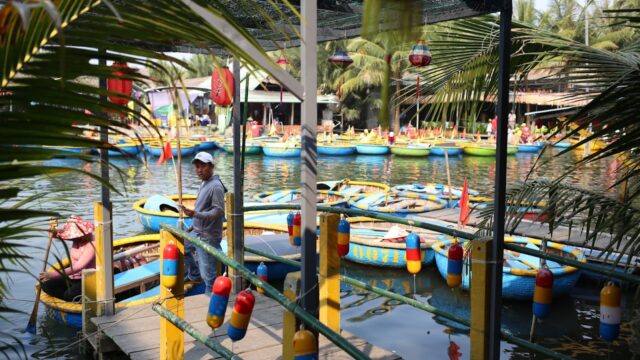 Colorful round boats float on a calm river in Hoi An near a dock with palm trees and people sitting and standing nearby.