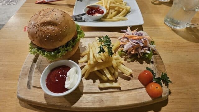 A burger, fries, salad, and tomatoes on a wooden plate evoke Hoi An café vibes, with drinks and another plate of fries across the table.