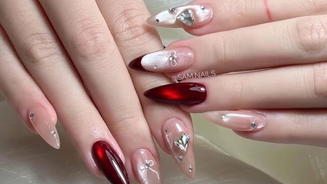 Hands with almond-shaped nails, painted red and nude with silver heart, bow, and gems, rest on white fabric in Hoi An.