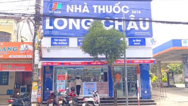 A pharmacy with a blue Nhà Thuốc Long Châu sign and motorbikes parked in front, situated on a bustling Hoi An city street.