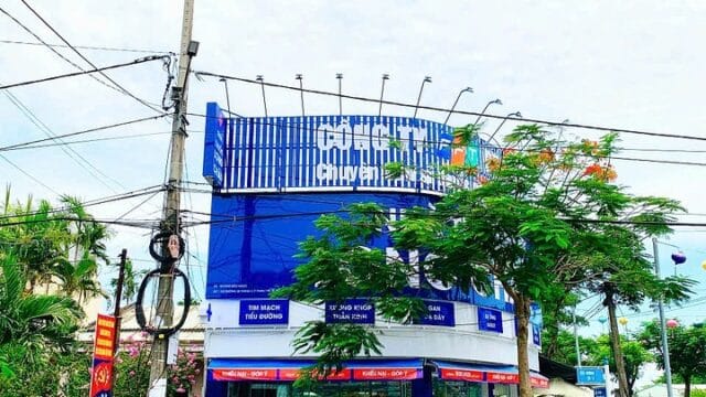 A blue and white pharmacy building on a street corner in Hoi An, with parked motorbikes and trees nearby under a cloudy sky.