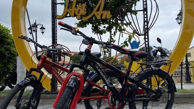 Three bicycles are parked in front of a yellow Hoi An sign, with blue lanterns and palm trees in the vibrant Hoi An backdrop.