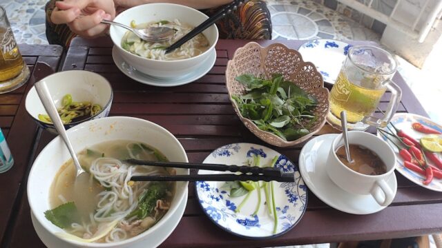 Two bowls of Hoi An noodle soup, herbs, sauces, tea, and coffee on a wooden table with two people dining.
