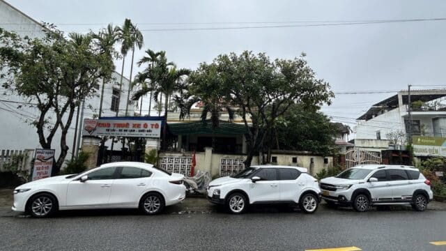Three white cars are parked in a row on a wet street in Hoi An, with trees and signage visible on a cloudy day.