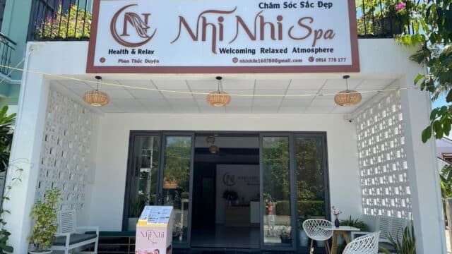 Front view of Nhi Nhi Spa in Hoi An with white chairs, plants, and a signboard above the entrance on a sunny day.