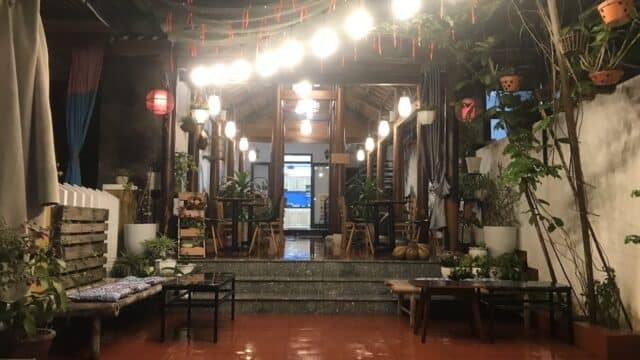 Cozy, plant-filled Hoi An courtyard with hanging lights, wooden benches, red tile floor, and tables leading into a warmly lit interior.