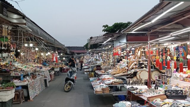 A busy open-air market in Hoi An with colorful stalls, goods, and a person riding a scooter down the aisle.