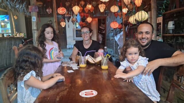 A family enjoys playing cards and drinking juice at a cozy Hoi An restaurant, sitting around a wooden table under warm lantern light.