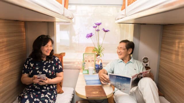 Two people sit on opposite bunk beds in a cozy train cabin, smiling and reading magazines on their journey to Hoi An.