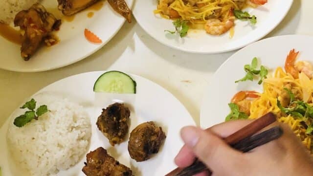 A hand uses chopsticks to pick meat from plates of rice, noodles, shrimp, and chicken wings in Hoi An.
