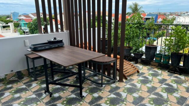 A wooden table and grill sit on a tiled balcony in Hoi An, with potted plants and a cityscape view in the background.