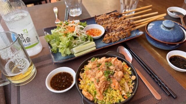 A table set with grilled meat skewers, fried rice, fresh salad, dipping sauces, and a bottle of soju in charming Hoi An.