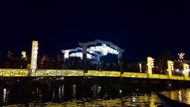 A brightly lit bridge in Hoi An with decorative lights reflected in the water at night, crowded with people.