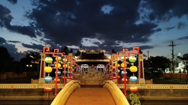 A brightly lit, ornate bridge in Hoi An with colorful lanterns at dusk leads to a temple-like structure under a cloudy sky.